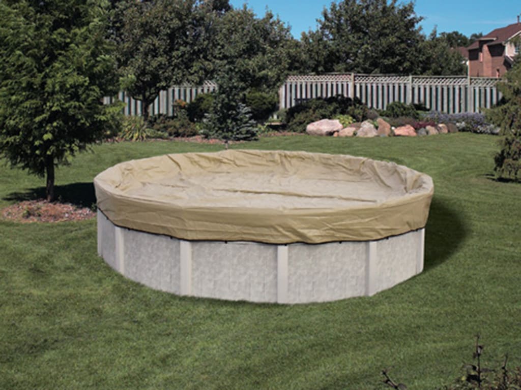 A Pool Goods HPI Armor Kote 30' Round Winter Cover (4' overlap, Tan/Blue) protects an above-ground pool on a grassy lawn, surrounded by trees, shrubs, and a white fence for reliable winter protection.