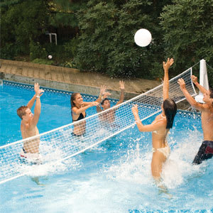 Five people play volleyball in a pool, jumping and reaching for the ball over a Swimline PoolJam In-Ground Pool Basketball & Volleyball Game Combo (9190) net, with trees and pavement visible in the background.
