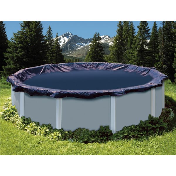 The Swimline Deluxe 18' Round Winter Cover (PCO822) with a 4' overlap in blue/black by Swimline protects your above-ground pool all winter, shown surrounded by grass, plants, and trees with snowy mountains in the distance.