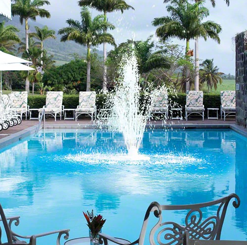 A swimming pool featuring the Swimline 12" Grecian Triple Tier Pool Fountain (8597) at its center, surrounded by lounge chairs, palm trees, and a scenic mountain view in the background.