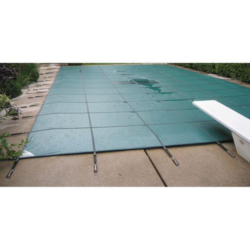 A green safety cover secured over an in-ground swimming pool with metal anchors, surrounded by a concrete deck and nearby plants.