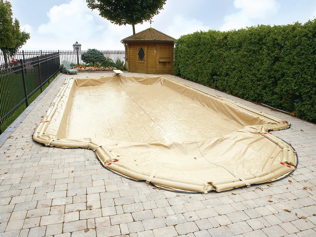 A rectangular swimming pool is covered with a green safety cover secured with straps and springs to the surrounding concrete deck.